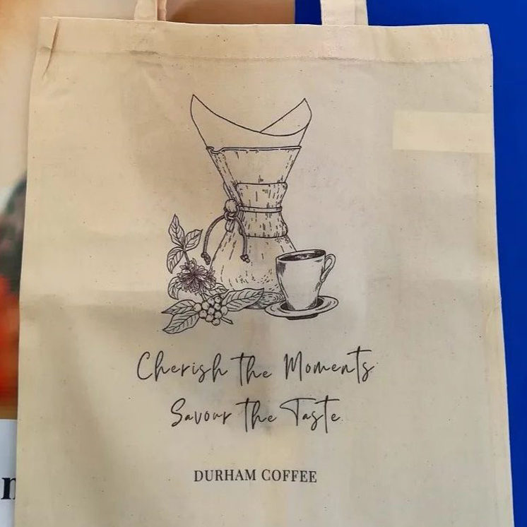 A natural coloured tote bag from Durham Coffee Roasters with the slogan 'Cherish the moments - Savour the taste' and a drawing of a chemex, coffee cup and coffee plant.